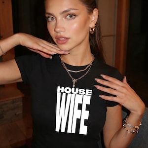 May include: Black crew neck t-shirt with the text "HOUSE WIFE" in bold white letters. The person is wearing gold jewellery, including a necklace and earrings.