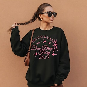 May include: Black crewneck sweatshirt with the text "The Nutcracker Dew Drop Fairy 2025" in pink. The design includes a ballerina and star accents. The sweatshirt is a casual style.