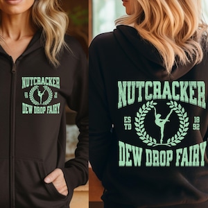 May include: A dark brown zip-up hoodie with the text "NUTCRACKER" above "DEW DROP FAIRY" in a mint green color. A ballerina graphic is in the center. The hoodie is a comfortable, casual garment.