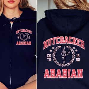 May include: Navy blue zip-up hoodie with the text "NUTCRACKER ARABIAN" in a distressed, collegiate-style font. The design includes a ballerina silhouette within a laurel wreath. The hoodie has a drawstring hood and a front zipper.