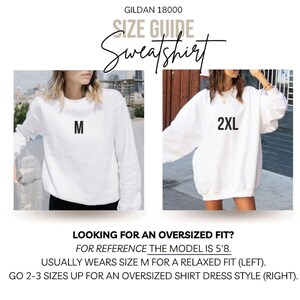 May include: White Gildan sweatshirt size guide. The image shows two women wearing white sweatshirts, one labeled "M" and the other "2XL". Text at the bottom reads "Looking for an oversized fit?" and provides sizing guidance.