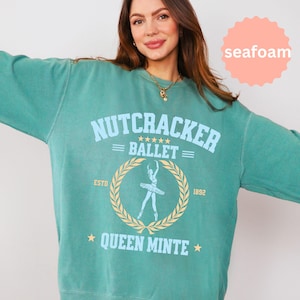 May include: A seafoam green crewneck sweatshirt featuring a graphic print with the text Nutcracker Ballet, Estd 1892, and Queen Minte. The design includes a central illustration of a ballerina inside a laurel wreath, accented by stars.