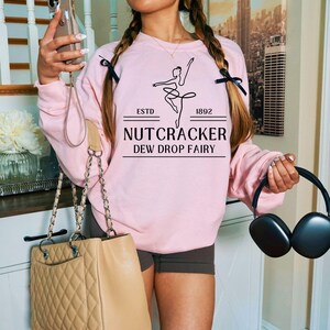 Può includere: Una felpa rosa chiaro con la scritta "NUTCRACKER DEW DROP FAIRY" e una grafica di ballerina. La felpa ha anche la scritta "ESTD 1892". La felpa è un modello girocollo.