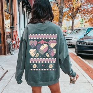 Puede incluir: Una sudadera de color verde azulado con un diseño de corazón de patchwork en la espalda. El diseño incluye corazones en varios patrones y colores, con detalles florales y geométricos. La sudadera tiene un aspecto de lavado vintage.