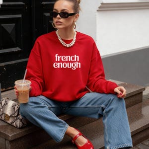 May include: A red sweatshirt with the text "french enough" in white. The person is wearing blue jeans, red shoes, black sunglasses, and a pearl necklace. They are holding a clear plastic cup with a straw.