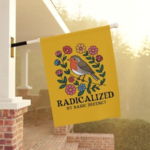 Puede incluir: Una bandera amarilla con una ilustración de un pájaro rodeado de flores. El pájaro tiene el pecho rojo y está posado en una rama. El texto "RADICALIZED BY BASIC DECENCY" está impreso en negro.