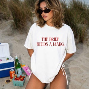 以下が含まれることがあります： 赤い文字で「THE BRIDE NEEDS A MARG」と書かれた白いTシャツ。ビーチで、クーラーボックス、飲み物、軽食が背景に写っています。サングラスをかけています。