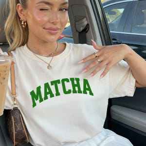 Op de afbeelding: Cr&egrave;mekleurig T-shirt met het woord "MATCHA" in vette groene letters. De persoon draagt gouden oorringen en meerdere gouden kettingen. De persoon houdt een drankje vast in een doorzichtige beker met een bruine schoudertas.
