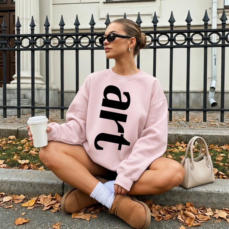 May include: A light pink sweatshirt with the word "art" in large black letters. The person is wearing sunglasses, white socks, and brown boots. A small beige handbag sits nearby.