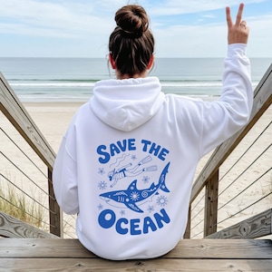 May include: White hoodie with a blue graphic on the back that reads "Save the Ocean". The design features a diver, a shark, and floral elements. The person is sitting on wooden steps with the sea in the background.