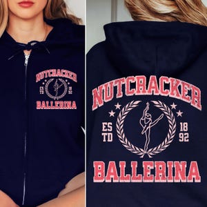 May include: Navy blue zip-up hoodie with a Nutcracker Ballerina design. The front features the text "Nutcracker Ballerina" in a distressed red font, with a ballerina graphic. The back has the same design. A stylish and comfortable garment.