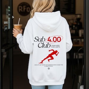 Puede incluir: Sudadera blanca con el texto "Sub 4.00 Club" en negro y rojo, junto con un gráfico rojo de un corredor. La sudadera también presenta las frases "Train Hard. Push Limits." y "An elite network of runners who chase the 4-minute mile."