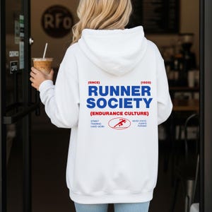 Puede incluir: Sudadera con capucha blanca con el texto "RUNNER SOCIETY" en azul, con detalles en rojo. La sudadera incluye el texto "(ENDURANCE CULTURE)" y un gráfico de una figura corriendo. La sudadera también tiene el texto "STREET TRAINING HARD WORK" y "NEVER STATIC ALWAYS PUSHING".