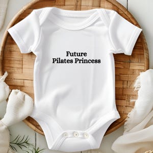 May include: White baby bodysuit featuring the text "Future Pilates Princess" in black. The garment has short sleeves and poppers at the bottom. The bodysuit is on a woven, circular tray with a white background.