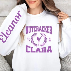 May include: White sweatshirt with the name "Eleanor" in purple script on the left sleeve. The front features the words "Nutcracker Clara" in purple, with a ballerina graphic and the year "1892".