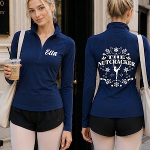 May include: A navy blue long-sleeve quarter-zip pullover featuring the name Ella in white script on the front left chest. The back displays a white graphic of a ballet dancer surrounded by snowflakes and the text The Nutcracker.