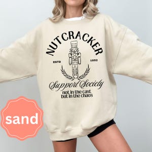 May include: Sand-colored sweatshirt with a black graphic of a nutcracker, the text "NUTCRACKER" arched above the image, and "Support Society" below. The sweatshirt also features the text "ESTD 1892" and "not in the cast but in the chaos".
