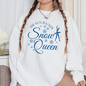 May include: White sweatshirt featuring the text "The Nutcracker Snow Queen" in blue script, with snowflake and ballerina graphics. A long-sleeved top suitable for winter or festive occasions.