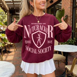 May include: A maroon long-sleeve crewneck sweatshirt featuring a light pink graphic print that reads ESTABLISHED 1892 NUTCRACKER SOCIAL SOCIETY with a central shield emblem containing a silhouette of a ballet dancer in a tutu.