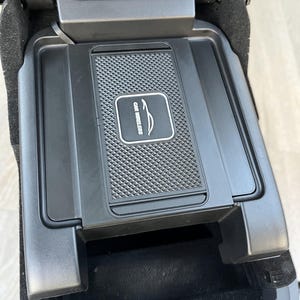 May include: A black car wireless charging pad with a textured surface and the text "CAR WIRELESS". The pad is integrated into a car's centre console, featuring a storage compartment below.