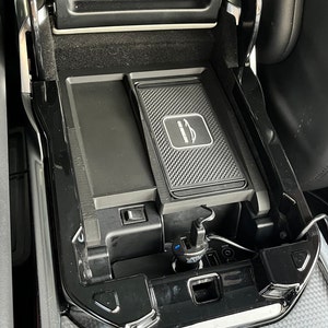 Può includere: Un interno di un'auto nero con un tappetino di ricarica wireless e una porta USB. Il tappetino di ricarica ha una superficie in gomma nera con un piccolo logo di un'auto al centro. La porta USB si trova sotto il tappetino di ricarica e ha un connettore blu e nero.