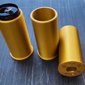 May include: Three gold-colored metal can holders. The can holders are shaped like bullets. One can holder has a black can inside. The can holder with the can inside has the top removed. The can holder on the right has the text "44 MAG" printed on the side.