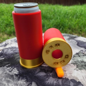 May include: Two red and gold shotgun shell-shaped can coolers. The top of the cooler is open and the bottom of the cooler has a gold band around it. The cooler is sitting on a camouflage-patterned surface.