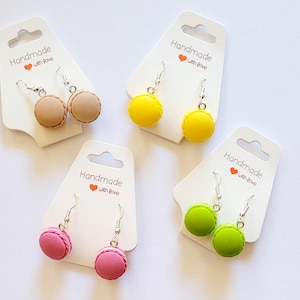 Könnte beinhalten: Vier Paar Hängeohrringe mit Miniatur-Macarons in verschiedenen Farben, darunter rosa, gelb, grün und beige. Jedes Paar wird auf einer weißen Karte mit dem Text "Handmade with love" präsentiert.