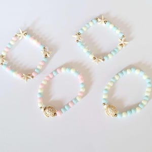 Könnte beinhalten: Vier farbige Perlenarmbänder mit Charms. Zwei Armbänder haben einen Seestern-Anhänger und zwei einen Schildkröten-Anhänger. Die Armbänder sind aus pastellblauen, rosa und weißen Perlen gefertigt.
