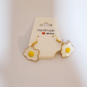 Könnte beinhalten: Ein Paar handgefertigte, goldfarbene Ohrringe in Form von Spiegeleiern. Jeder Ohrring hat ein weißes Emaille-Eiweiß mit einem gelben Eigelb in der Mitte und einem goldfarbenen Rand. Die Ohrringe werden auf einer cremefarbenen Karte mit dem Text "Handmade with love" präsentiert.