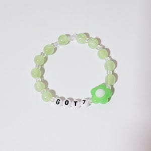 Könnte beinhalten: Ein grünes Perlenarmband mit einem grünen Blumenanhänger und weißen Buchstabenperlen, die "GOT7" buchstabieren.