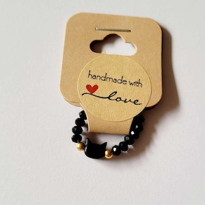 Könnte beinhalten: Schwarzes Perlenarmband mit einem schwarzen Katzenanhänger und goldenen Perlen. Das Armband befindet sich auf einer braunen Karte mit dem Text "Handgefertigt mit Liebe" und einem roten Herzen.