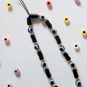 Könnte beinhalten: Schwarzer Handy-Anhänger mit blauen Evil-Eye-Perlen und goldenen Akzenten. Der Anhänger ist an einer schwarzen Kordel befestigt und enthält bunte, verstreute Perlen.