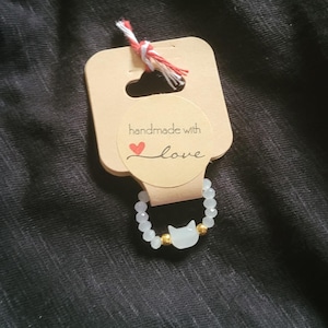 Könnte beinhalten: Weißes Perlenarmband mit einem goldenen Katzenanhänger. Das Armband befindet sich auf einem braunen Etikett mit dem Text "handgefertigt mit Liebe" und einem roten Herzen.