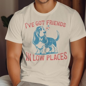 This retro dog shirt features a vintage dog tee design of a basset hound standing proudly, tail high, and gazing to the side. Text reads “I’ve got friends” above and “in low places” below. A distressed texture adds charm to the shirt.