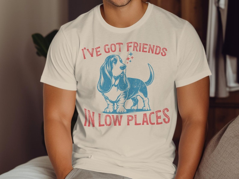 This retro dog shirt features a vintage dog tee design of a basset hound standing proudly, tail high, and gazing to the side. Text reads “I’ve got friends” above and “in low places” below. A distressed texture adds charm to the shirt.