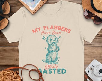 Puede incluir: Una camiseta beige con un gráfico de una nutria con el texto "My flabbers have been otterlly gasted" en azul y rosa.