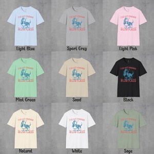 May include: A set of ten t-shirts in various colors featuring a graphic of a dog with the text "I've got friends in low places". The colors include light blue, sport grey, light pink, mint green, sand, black, natural, white, and sage.