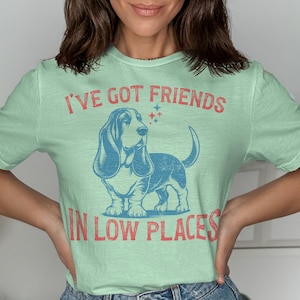 This retro dog shirt features a vintage dog tee design of a basset hound standing proudly, tail high, and gazing to the side. Text reads “I’ve got friends” above and “in low places” below. A distressed texture adds charm to the shirt.