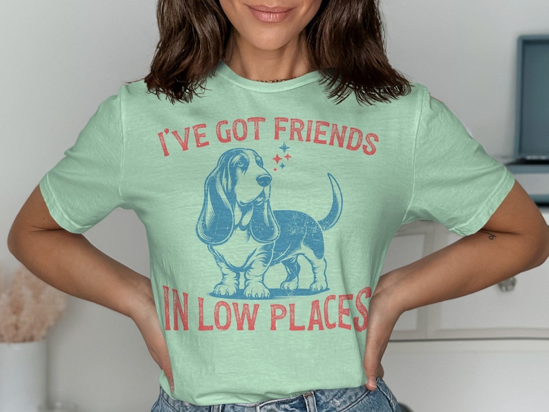 This retro dog shirt features a vintage dog tee design of a basset hound standing proudly, tail high, and gazing to the side. Text reads “I’ve got friends” above and “in low places” below. A distressed texture adds charm to the shirt.