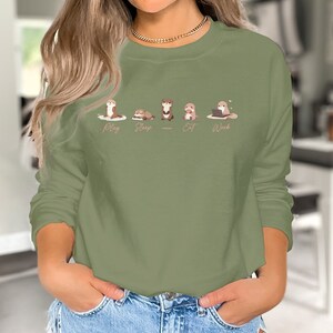 Pode incluir: Uma camisa de moletom verde com um gr&aacute;fico de cinco lontras em diferentes poses. As lontras s&atilde;o rotuladas como "Play", "Sleep", "Eat", "Work".