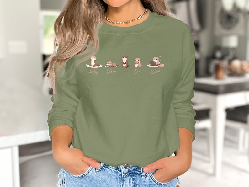 Pode incluir: Uma camisa de moletom verde com um gr&aacute;fico de cinco lontras em diferentes poses. As lontras s&atilde;o rotuladas como "Play", "Sleep", "Eat", "Work".