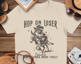 May include: A beige t-shirt with a black and white illustration of a raccoon riding a donkey. The text "Hop on Loser Trouble Ain't Gonna Make Itself" is printed on the shirt.
