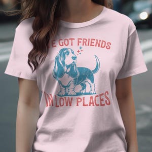 This retro dog shirt features a vintage dog tee design of a basset hound standing proudly, tail high, and gazing to the side. Text reads “I’ve got friends” above and “in low places” below. A distressed texture adds charm to the shirt.