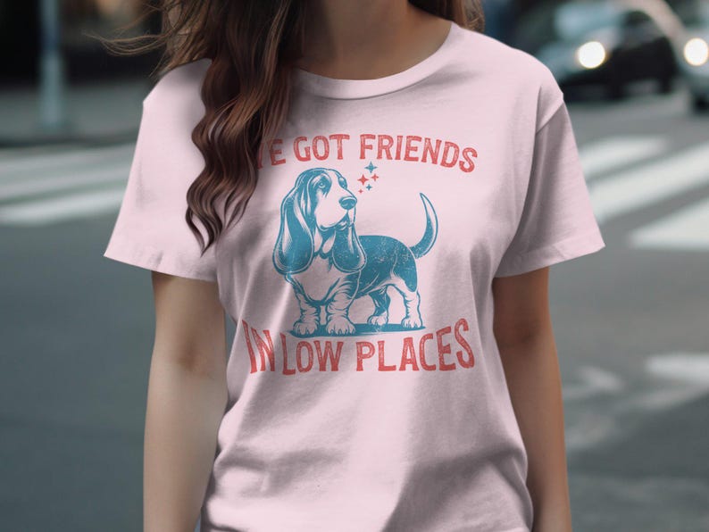 This retro dog shirt features a vintage dog tee design of a basset hound standing proudly, tail high, and gazing to the side. Text reads “I’ve got friends” above and “in low places” below. A distressed texture adds charm to the shirt.
