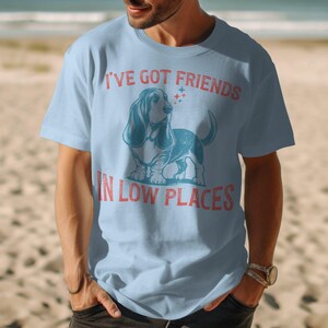 This retro dog shirt features a vintage dog tee design of a basset hound standing proudly, tail high, and gazing to the side. Text reads “I’ve got friends” above and “in low places” below. A distressed texture adds charm to the shirt.