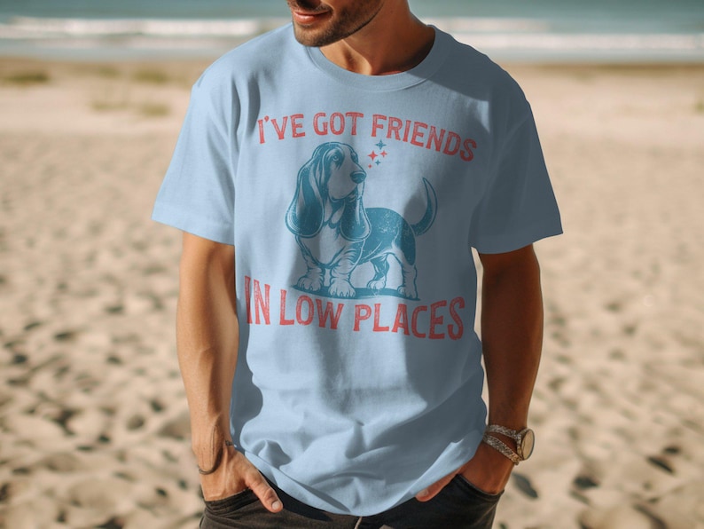 This retro dog shirt features a vintage dog tee design of a basset hound standing proudly, tail high, and gazing to the side. Text reads “I’ve got friends” above and “in low places” below. A distressed texture adds charm to the shirt.