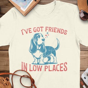 This retro dog shirt features a vintage dog tee design of a basset hound standing proudly, tail high, and gazing to the side. Text reads “I’ve got friends” above and “in low places” below. A distressed texture adds charm to the shirt.