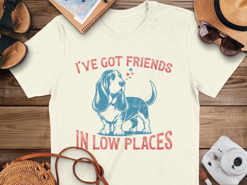 This retro dog shirt features a vintage dog tee design of a basset hound standing proudly, tail high, and gazing to the side. Text reads “I’ve got friends” above and “in low places” below. A distressed texture adds charm to the shirt.