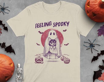 May include: Cream-colored t-shirt with a Halloween-themed graphic. The design features a ghost holding a small dog, with the words "FEELING SPOOKY" above. Bats, pumpkins, and a red moon complete the spooky scene.